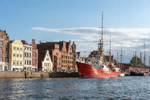 Blick über die Trave auf das andere Ufer, die Straße und die Häuser der Altstadt in Lübeck. Am gegenüberliegenden Ufer liegt das Segelschiff Fehmarnbelt.