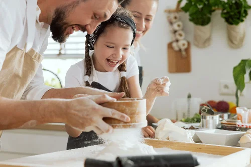 Nahaufnahme der neugierigen jungen Tochter, die Papa mit gebärtem Siebmehl hilft, hausgemachten Teig für die heimische Pizza vorzubereiten. Im Hintergrund die lächelnde Mutter.