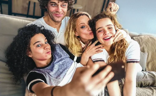 Familie, die gemeinsam auf einem Sofa sitzt und lachend in ein Handy schaut. Foto: filadendron, iStock.com