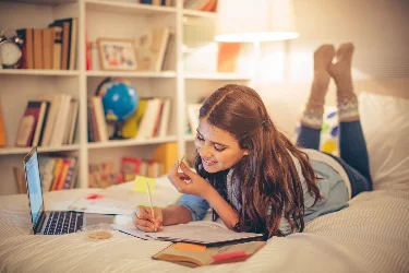 Ein Mädchen liegt bei gemütlichem Lampenlich auf seinem Bett, ein Laptop neben sich und schreibt in ein Heft. Foto: svetikd, iStock.com
